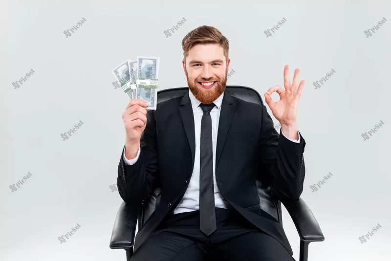 happy smiling business man black suit holding money one hand showing ok sign sitting armchair isolated gray background / مرد تاجر خندان شاد کت و شلوار مشکی و پول دلار در دست روی صندلی راحتی نشسته سود از سرمایه گذاری مالی