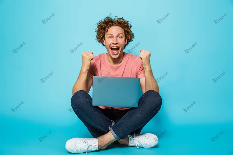 image caucasian happy man with brown curly hair rejoicing clenching fists while sitting floor with legs crossed / تصویر مرد شاد قفقازی با موهای مجعد قهوه ای در حالی که مشت های گره کرده و روی زمین با لپ تاپ نشسته است