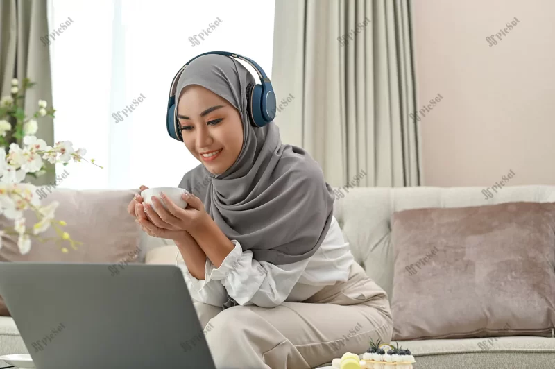 relaxed asian muslim woman wearing hijab headphones is watching movie her laptop / زن مسلمان آسیایی آرام با حجاب وهدفون در حال تماشای فیلم در لپ تاپ با فنجان چای