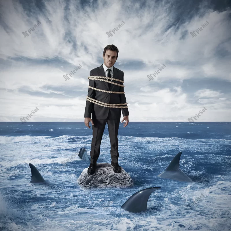 risk business / مرد با دستان بسته روی صخره ای در دریای پر از کوسه مفهوم ریسک در کسب و کار